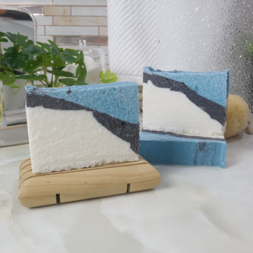 A selection of soaps are displayed on a bathroom countertop along with a plant and hand mirror.   One soap stands on a wooden soap dish to the left of the image.  To the right and just behind, another soap lays flat with another soap standing upright on it.  All soaps have a diagonal colour pattern that has a large white  area on the lower left, which flows into a black narrow line and finished with a bright blue strip.