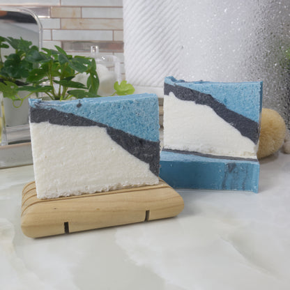 A selection of soaps are displayed on a bathroom countertop along with a plant and hand mirror.   One soap stands on a wooden soap dish to the left of the image.  To the right and just behind, another soap lays flat with another soap standing upright on it.  All soaps have a diagonal colour pattern that has a large white  area on the lower left, which flows into a black narrow line and finished with a bright blue strip.