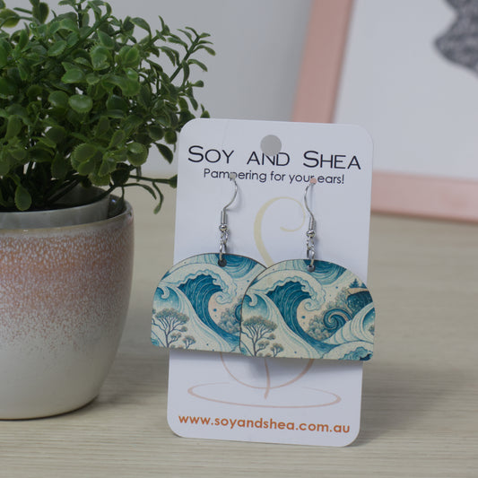 Ocean Waves Earrings by Soy and Shea are displayed on a branded card beside a green potted plant, set on a light-colored surface.  The earrings are a solid arch shape featuring a sublimated design of blue ocean waves
