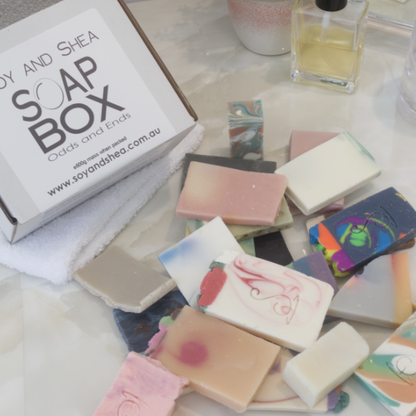 A selection of soap pieces lay scattered  across a bathroom counter next to a white box labelled SOAP BOX ODDS AND ENDS.  The soaps are various shapes and colours.