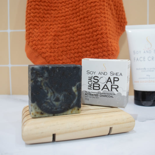 A small square shaped bar of soap along with its packaging stand on a wooden soap dish on a white surgace agains a tile background.  A towel and tube of FACE CREAM can be seen in the background.   The soap is black with wisps of pale green and flecks of brown