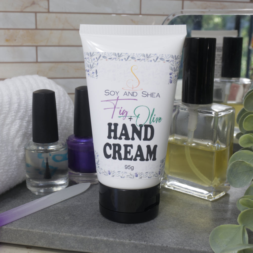 A White tube with black lid stands on a grey stone surface surrounded by two bottles of nail polish, a nail file, bottle of perfume and a mirror.  The tube has a decorative product label identifying it as Fig and Olive Hand Cream and features a decorative pattern of olive leaves and figs.
