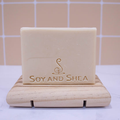 A cream bar soap stands on a wooden soap dish on white surface with a faded peach tile background. 