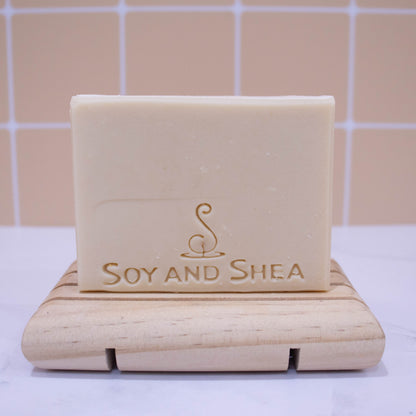 A bar of soap stands on a wooden soap dish on a white surface with peach tile background.  The soap is a solid cream colour and stamped with logo and SOY AND SHEA