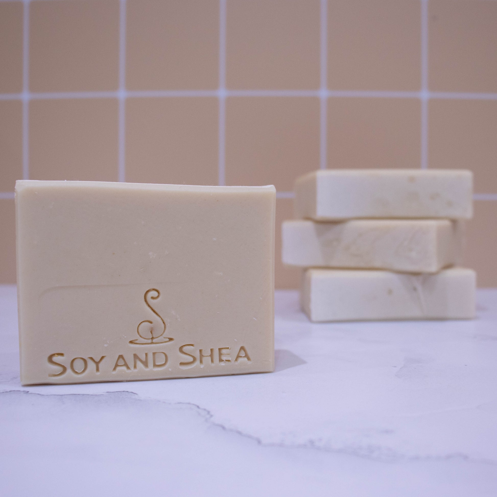 A collection of soaps sit on a white marble surface against a pale peach tile background.  In the background, three soaps lay flat but stacked upon each other revealing a textured top.   A fourth soap appears in the foreground showing a solid cream soap embossed with SOY AND SHEA