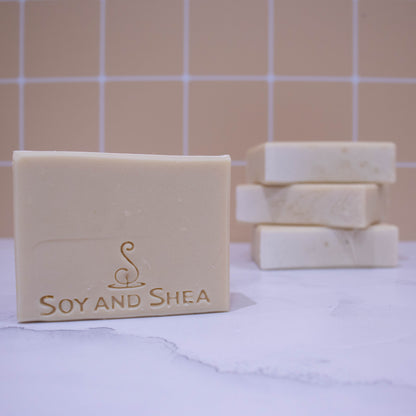 A collection of soaps sit on a white marble surface against a pale peach tile background.  In the background, three soaps lay flat but stacked upon each other revealing a textured top.   A fourth soap appears in the foreground showing a solid cream soap embossed with SOY AND SHEA