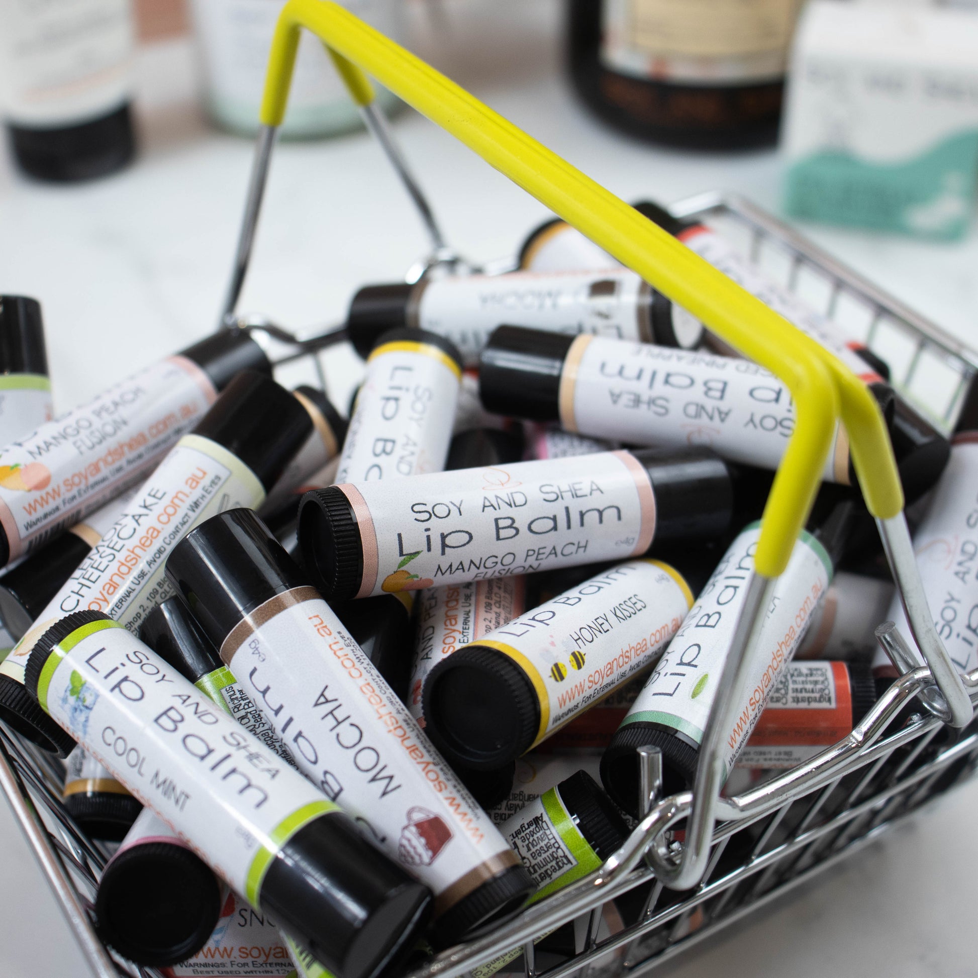 A small metal  basket with yellow handles is filled with various flavoured lip balms.  Ths is set against a blurred background