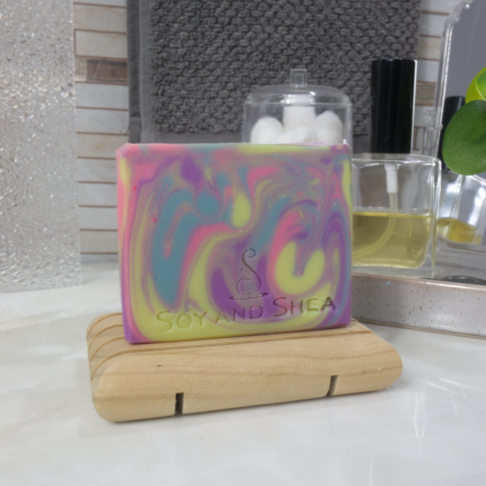 A rectangular bar of soap stands on a wooden soap rack on a bathroom countertop next to a bottle of perfume and jar of cotton balls.  The soap has a rainbow swirl of bright neon colours in Yellow, blue, pink and purple