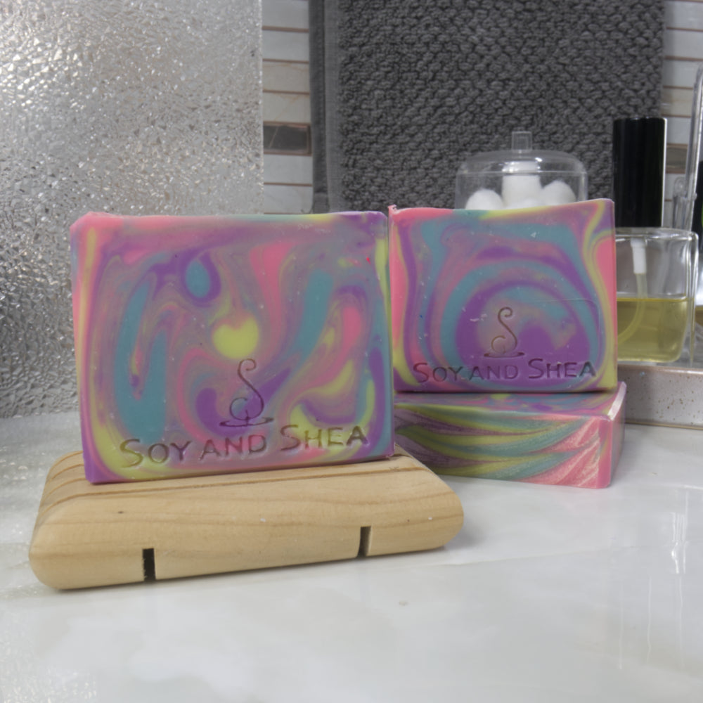 A selection of soaps are presented on a bathroom countertop. One soap stands upright on a wooden soap rack and to the back left, another lays flat on the bench with a third soap standing on top.   The soaps have bright swirling colurs of neon blue, pink, yellow and purple