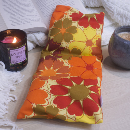 A rectangular heat pack with an orange, yellow, and red floral pattern, displayed next to a lit candle and an open book.