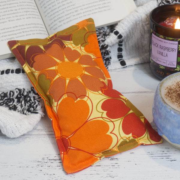 A rectangular mini heat pack with a vibrant orange and red floral pattern, displayed on a wooden surface alongside a lit candle and an open book.  The Mini Heat Pack is rectangular with a pale yellow background and bright orange and red flowers with heart shaped petals.
