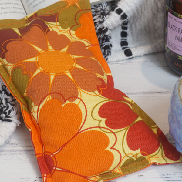 An Orange Bloom Mini Heat pack rests on a wooden bench, propped up on a black and white blanket.  The image shows a close up of the fabric design which is a pale yellow background with large orange and red stylised flowers with heart shaped petals.