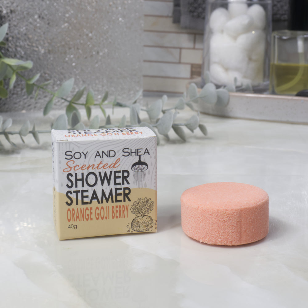 A boxed scented shower steamer and the steamer itself on a bathroom counter. The box reads 'Soy and Shea Scented Shower Steamer Orange Goji Berry', and the round orange steamer sits beside it.
