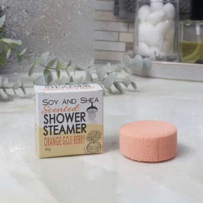 A boxed scented shower steamer and the steamer itself on a bathroom counter. The box reads 'Soy and Shea Scented Shower Steamer Orange Goji Berry', and the round orange steamer sits beside it.