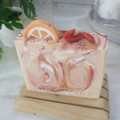 An soap bar stands on a wooden soap dish on a bathroom countertop with a plant and stone tray.   The soap is pale orange with swirls of bright orange, red and white. The image focuses on the top of the soap which features soap decorations of an orange slice and some red berries and some red biodegradable glitter