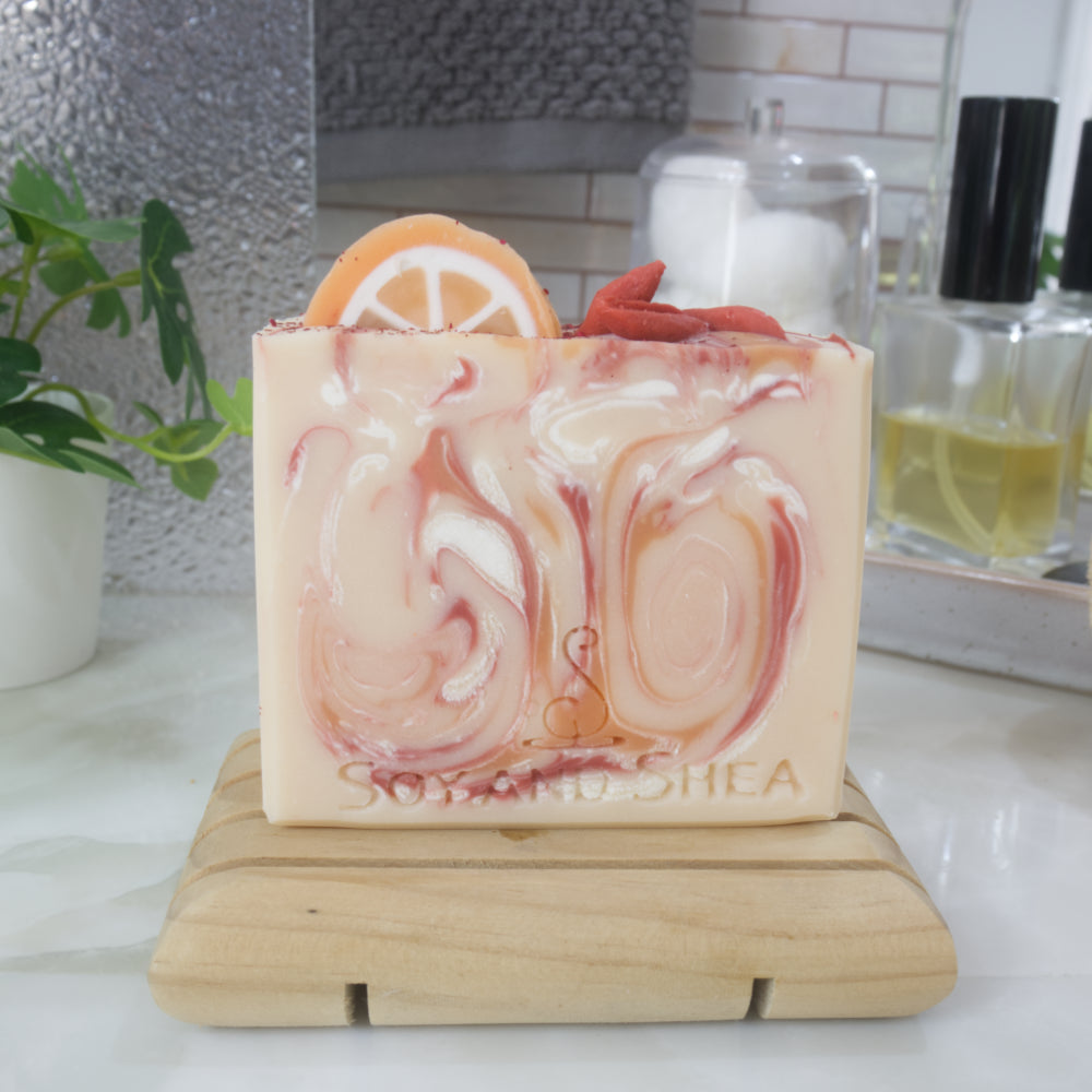 An soap bar stands on a wooden soap dish on a bathroom countertop surrounded by perfume bottle, jar of cotton and a plant.   The soap is pale orange with swirls of bright orange, red and white.  The top has soap decorations of an orange slice and some red berries.