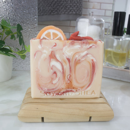 An soap bar stands on a wooden soap dish on a bathroom countertop surrounded by perfume bottle, jar of cotton and a plant.   The soap is pale orange with swirls of bright orange, red and white.  The top has soap decorations of an orange slice and some red berries.