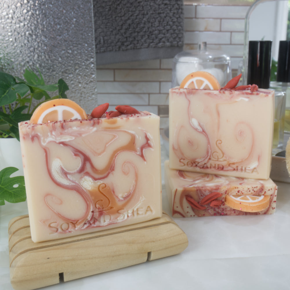 A collection of 3 soap bars are displayed on a bathroom countertop surrounded by perfume bottle, towel and plant.  One soap bar is standing on a wooden soap dish while another two are positioned behind with one lying flat to show the top of the soap and another standing on it.  All soaps are a pale orange with each having varying swirls of colour in bright orange, red and white.  The soaps feature soap decorations on the top made to look like half an orange slice in the along with goji berries