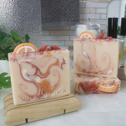 A collection of 3 soap bars are displayed on a bathroom countertop surrounded by perfume bottle, towel and plant.  One soap bar is standing on a wooden soap dish while another two are positioned behind with one lying flat to show the top of the soap and another standing on it.  All soaps are a pale orange with each having varying swirls of colour in bright orange, red and white.  The soaps feature soap decorations on the top made to look like half an orange slice in the along with goji berries