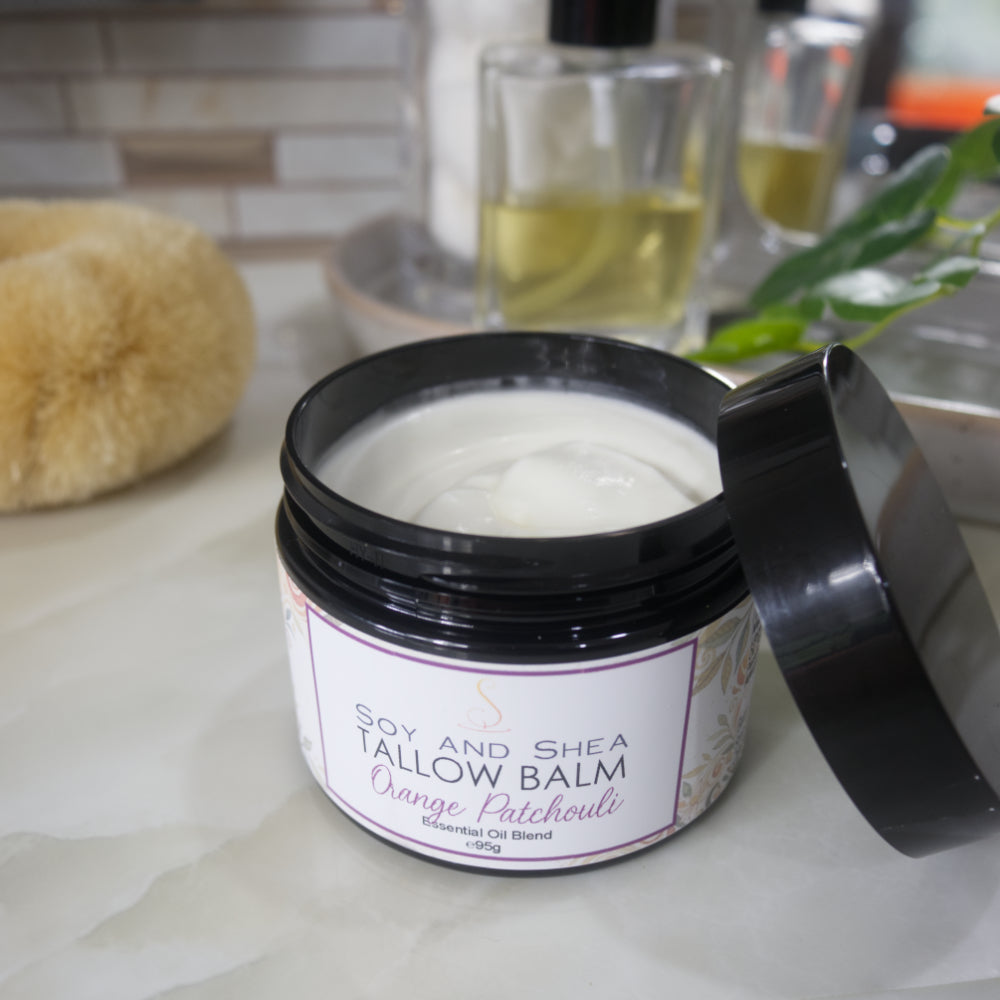 A black jar with a decorative label is displayed on a bathroom countertop.  The label identifies it as Tallow Balm in Orange and Patchouli.  The lid of the jar rests on the side of the jar revealing a white balm inside.