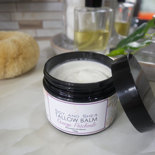 A black jar with a decorative label is displayed on a bathroom countertop.  The label identifies it as Tallow Balm in Orange and Patchouli.  The lid of the jar rests on the side of the jar revealing a white balm inside.
