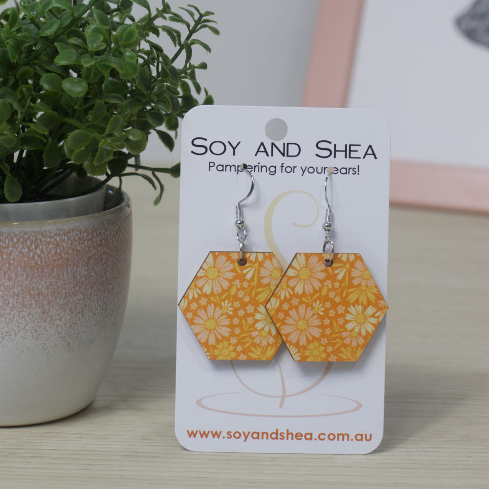Orange Petals Earrings by Soy and Shea—a pair of hexagonal, sublimated earrings with a yellow floral design on orange background—are displayed on a branded card beside a small potted plant on a light wooden surface.