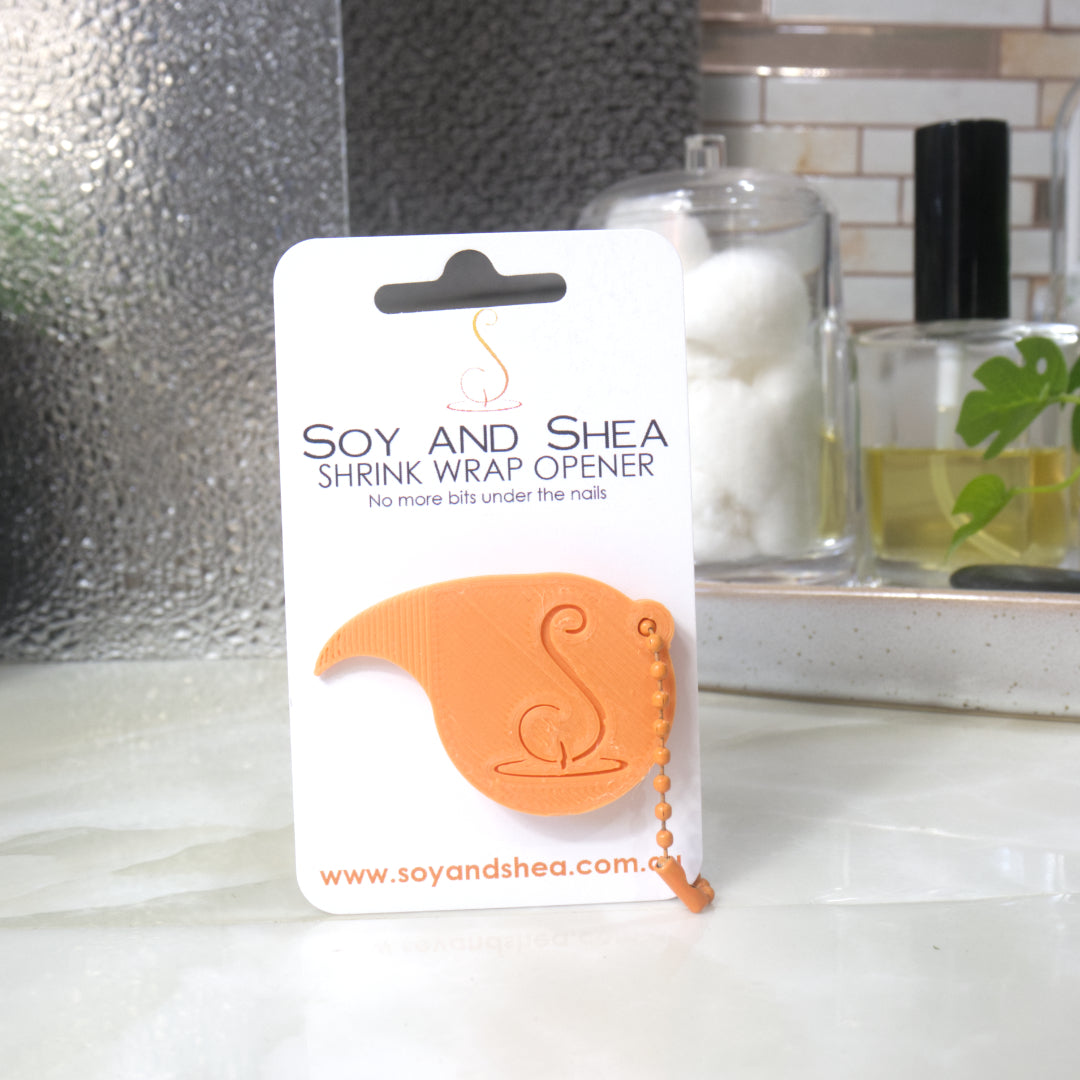 An Orange Shrinkwrap opener with matching ball and chain keyring is attached to a white card with product details.  The card is standing upright on a bathroom countertop with a bottle of perfume, a jar of cotton balls and towel in the background