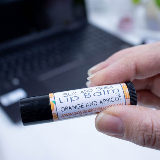 A hand holds a black tube of lip balm against a blurred background with an open laptop computer on a white surface.  The label has text LIP BALM ORANGE AND APRICOT on a white background with a pale orange border with a random pattern of orange and apricot drawings.