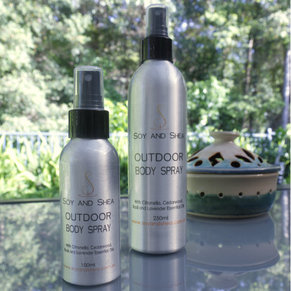 Two silver bottles labeled "Outdoor Body Spray" by Soy and Shea are placed on a glass table. One bottle is smaller, containing 100ml, while the other is larger at 250ml. In the background, there is a decorative ceramic item and lush greenery adds a natural touch behind them.