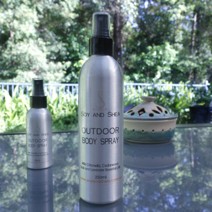 Two bottles of Soy and Shea's Outdoor Body Spray, enriched with essential oils, are displayed on a glass table in a tranquil outdoor setting. The larger bottle stands prominently in front, with the smaller one slightly obscured behind it. A decorative object enhances the charm of the background.