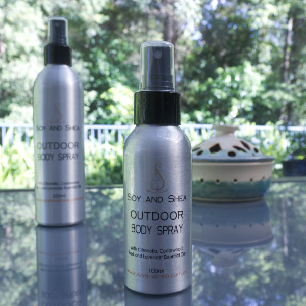 Two bottles of "Outdoor Body Spray" by Soy and Shea, featuring essential oils such as citronella, cedarwood, basil, and lavender, rest on a reflective outdoor surface. A decorative container is positioned in the background with trees visible in the distance.