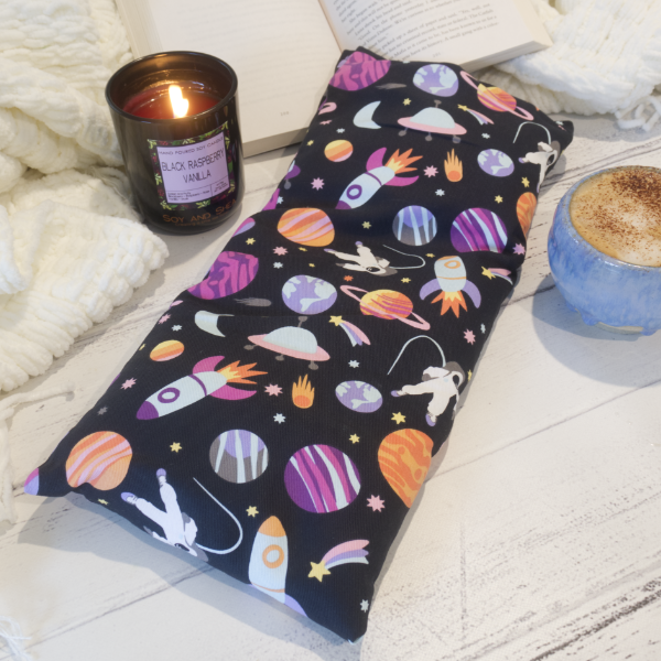 A long rectangular heat pack with a black background featuring colorful space-themed patterns, including planets and rockets, and a white astronaut. The heat pack is displayed next to a candle and a cup of coffee on a wooden surface with a white fluffy blanket in the background.