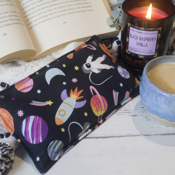 A mini rectangular heat pack rests against a black and white blanket and open book.  Next to it is a burning candle and filled coffee cup evoking a comfy scene.   The heat pack has a black fabric with illustrations of brightly coloured planets and stars along with a white astronaut.