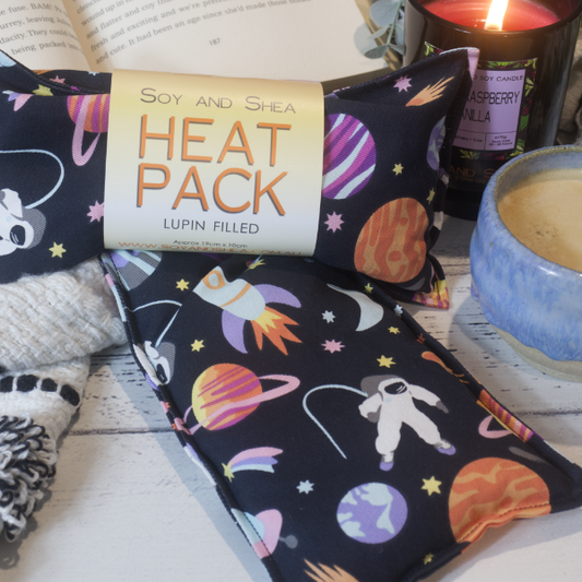 Two Mini Heat packs are stacked upon each other, one with a belly band label reading HEAT PACK LUPIN FIlled.   These are resting on a black and white blanket with open book, candle and coffee cup evoking a cosy feel.    The Heat packs have a black fabric with illustrations of various planet, a white astronaut, rocket and stars