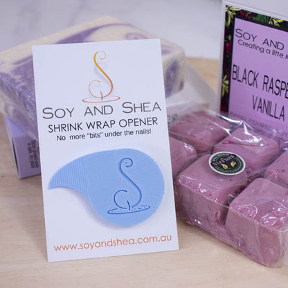 A white business-sized card branded SOY AND SHEA leans against a purple and white bar of soap and a large magenta coloured melt block both wrapped in a plastic film.   Additional text on the card reads SHRINK WRAP OPENER NO MORE BITS UNDER THE NAILS.   Stuck to the card is a teardrop shaped shrinkwrap opener that has the logomark embossed into with the point of the teardrop bent into a sharp point.  The opener is a light blue.