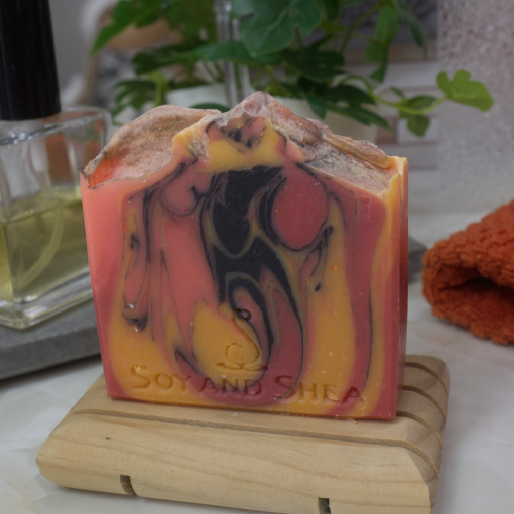 Colorful soap bar with marbled design on a wooden stand, with a plant and towel in the background.  The soap features colours of bright orange, yellow and black