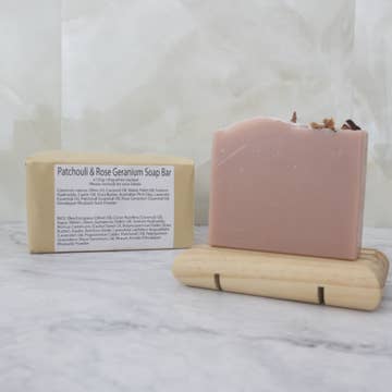 A dusky pink soap bar sits on a wooden stand with a brown paper package behind labeled 'Patchouli and Rose Geranuium Soap Bar' on a marble background