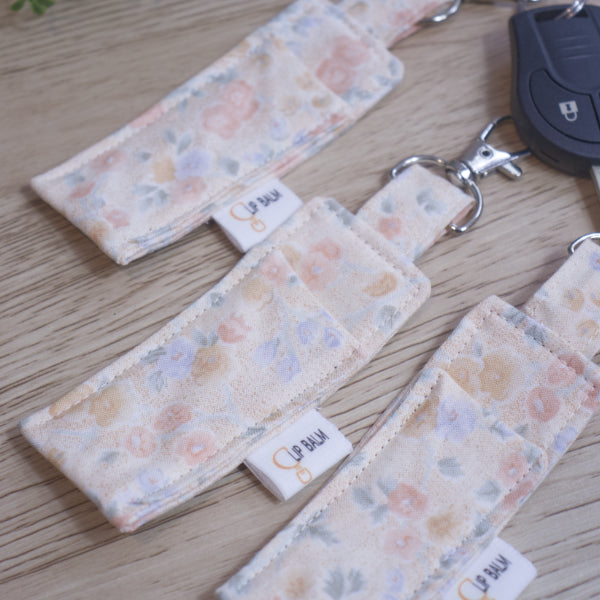 A selection of three lip balms showing variations in fabric design, lie on a wooden side next to a car key .  The Clip balms have a pale peach background and are covered in watercolour flowers in shades of yellow, blue and dark peach.
