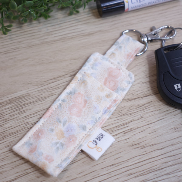 A Lip Balm holder lies on an wooden surface with lobster claw clasp attached to a car key.  A lip balm and greenery can be seen in the image.  The Clip Balm has a pale cream mottled background covered with simplistic watercolur flowers in peach, blue and yellow