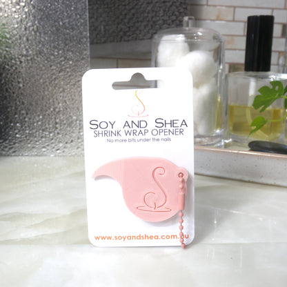 A peach Shrinkwrap opener with matching ball and chain keyring is attached to a white card with product details.  The card is standing upright on a bathroom countertop with a bottle of perfume, a jar of cotton balls and towel in the background