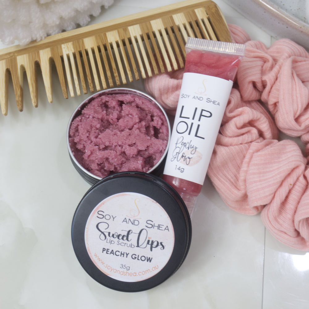 A small black tin with the lid removed revealing a dusky rose Lip Scrub sits on a marble tile surface next to a small, clear tube of peachy coloured Lip Oil.   This rests againsts a wooden comb and pink scrunchie. 