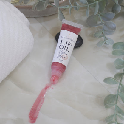 A tube of Lip Oil lies on a marble tile surface next to a white towel and some eucalyptus leaves.   The tube is clear with a white product label and filled with a peachy red Lip oil.  The cap is removed and a smear of Lip oil is on the bench showing the translucent effect it has