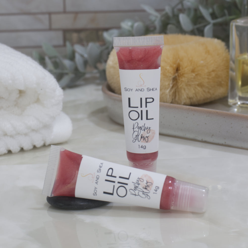 Two tubes of Lip Oil are presented on a bathroom countertop with a blurred background of towels, greenery and body brush.   One tube stands upright while the other lays flat in front of it.  The tubes are clear with a white product lable and are filled with a peachy red Lip oil.