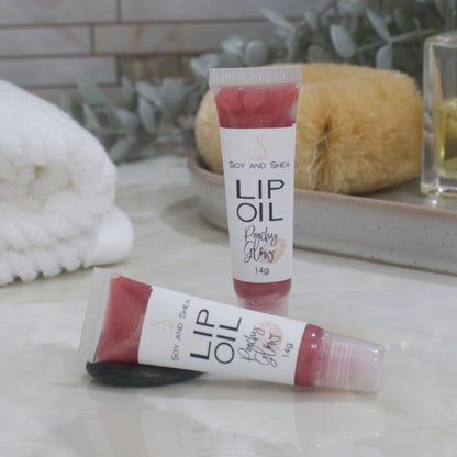 Two tubes of Lip Oil are presented on a bathroom countertop with a blurred background of towels, greenery and body brush.   One tube stands upright while the other lays flat in front of it.  The tubes are clear with a white product lable and are filled with a peachy red Lip oil.