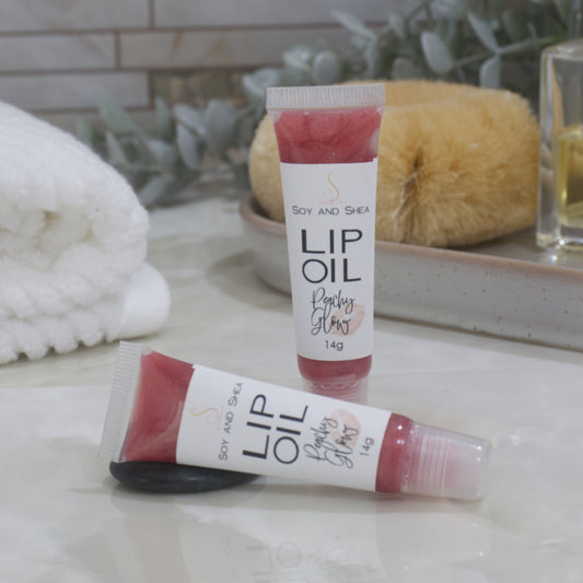 Two tubes of Lip Oil are presented on a bathroom countertop with a blurred background of towels, greenery and body brush.   One tube stands upright while the other lays flat in front of it.  The tubes are clear with a white product lable and are filled with a peachy red Lip oil.