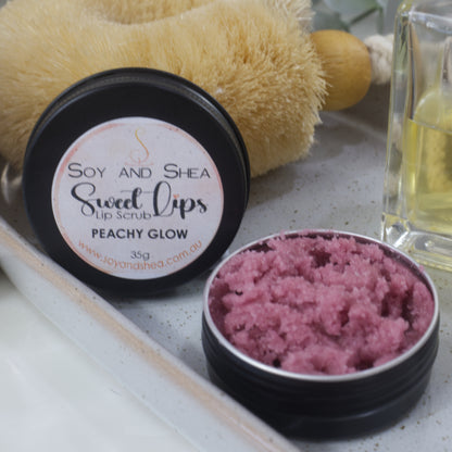 A round black tin sits on a stone tray along with a bottle of perfume and body brush.  The lid has been removed and is resting against the body brush revealing a dusky rose sugar scrub 
