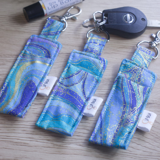 A selection of three clip balms showing variations in fabric design, lie on a wooden side with the middle Clip Balm attached to a car key.  A lip balm lays near-by along with some greenery.  The Clip balms have a wavy design in shades of blue with gold swirls highlighting the colours