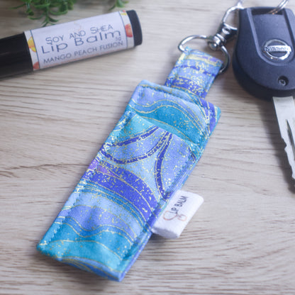 A lip balm holder lies on a wooden surface attached to a car key with a "lobster claw" clip.  To the side is a tube of lip balm. The Clip Balm has a wavy pattern in shades of teal, aqua, and  light and dark purple all with gold highlights