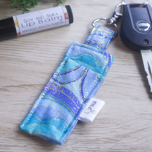 A lip balm holder lies on a wooden surface attached to a car key with a "lobster claw" clip.  To the side is a tube of lip balm. The Clip Balm has a wavy pattern in shades of teal, aqua, and  light and dark purple all with gold highlights