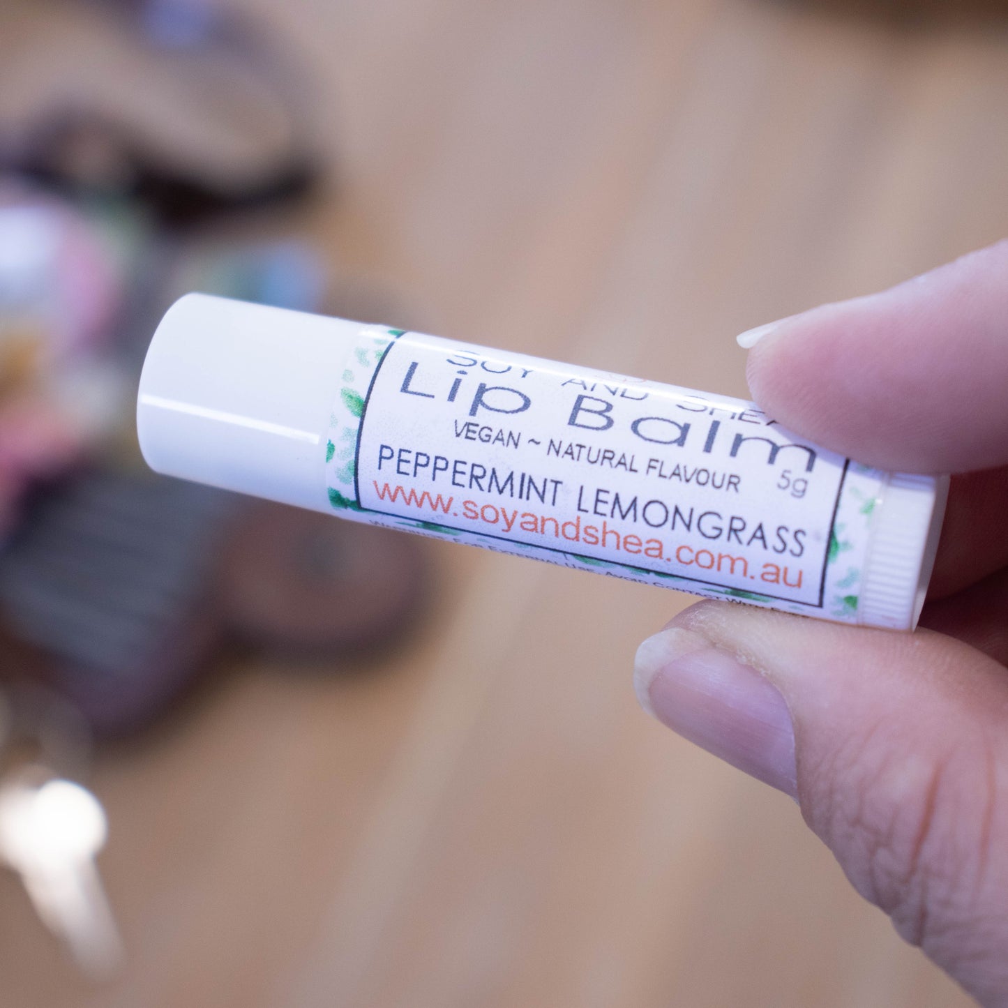 A hand holds a white tube labelled lip balm over a blurred image of an open handbag with contents spilling onto the table.  The lip balms features a white label with a random scattering of green leaves